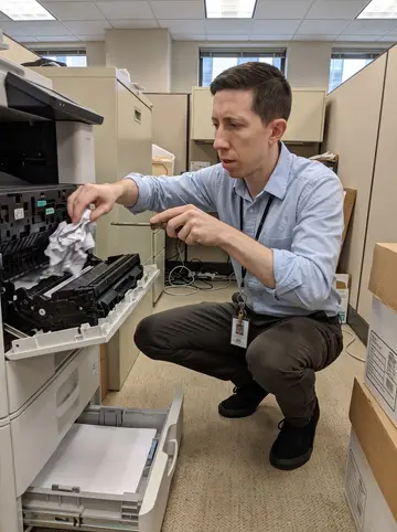 Conall, AI character: printer corner of a beige-carpeted office in downtown Pittsburgh, open toner drawer and a stack of paper…