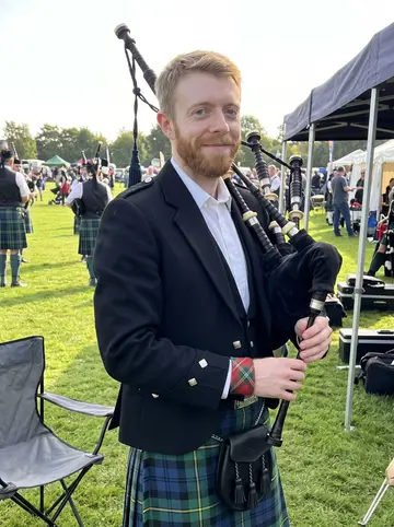 Arran, AI character: a late-summer Highland games field in Toronto, pipe band tents and a grass parade lane behind him