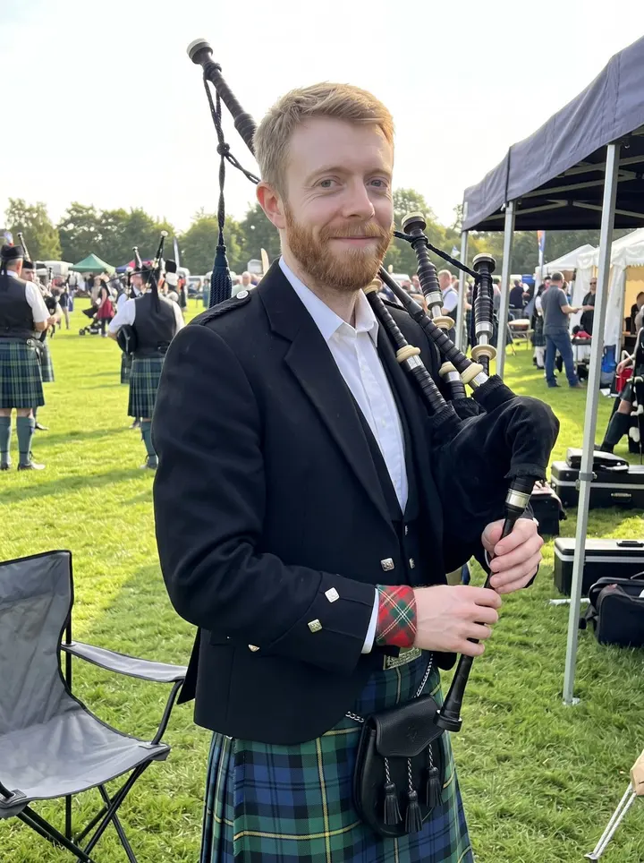 Arran, AI character: a late-summer Highland games field in Toronto, pipe band tents and a grass parade lane behind him