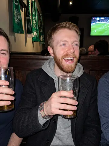 Arran, AI character: a busy Scottish pub in Toronto during a Celtic football match, dark wood booths and green scarves behind him