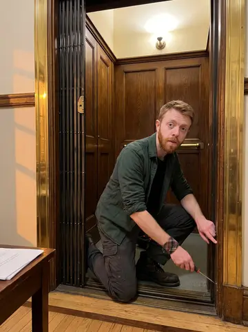 Arran, AI character: the lobby of his own older apartment building, beside a vintage wooden lift with brass trim and exposed…
