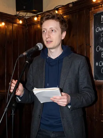 Fintan, AI character: a small old Dublin pub open-mic corner, dark wood wall and a chalkboard menu behind him