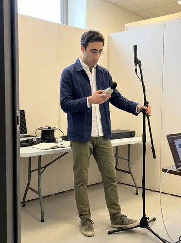 Gaetano, AI character: inside an acoustic test room at a renovation site in Turin, with pale acoustic panels and a laptop on a…