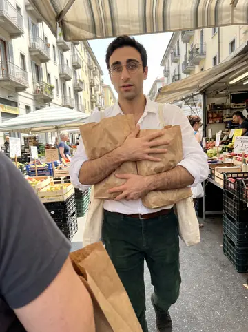 Gaetano, AI character: outside Porta Palazzo market on a Thursday grocery run for Signora Rosa, with produce stalls and apartment…