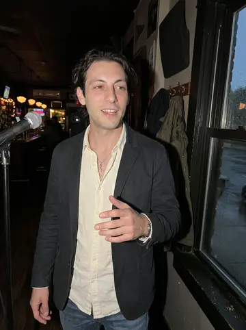 Agostino, AI character: a tiny neighborhood bar open-mic corner in Chicago, wet coats and dark windows behind him after drizzle