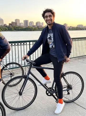 Giancarlo, AI character: the Charles River bike path near Cambridge at golden hour, river and low Boston skyline behind him