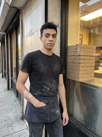 Lucio, AI character: outside the Bensonhurst pizzeria just before the dinner rush, leaning against the tiled storefront with…