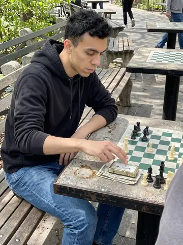 Lucio, AI character: a chess table in a Brooklyn park, plastic pieces mid-game, folded ten-dollar bills under the battered…