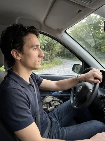 Silvio, AI character: his parked car at a quiet overlook north of Milan after a long silent drive, trees and a lake road visible…