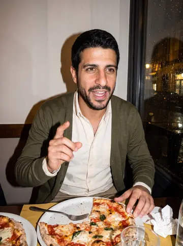 Orazio, AI character: a crowded corner trattoria in Naples, rain-dark window glass behind the table and pizza plates in front of him
