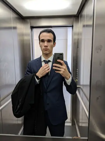 Oliviero, AI character: elevator mirror in a Milan office building before a morning client meeting, brushed metal walls and a…