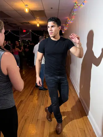 Tomás, AI character: a Little Havana salsa dance hall on a Thursday night, polished wood floor, colored string lights, a few…
