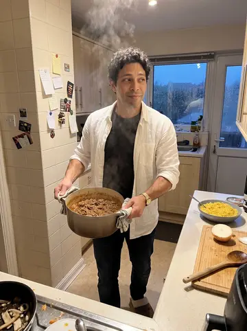 Jorge, AI character: his mother's kitchen above the garage, carrying a pot of ropa vieja toward the counter while rain beads on…