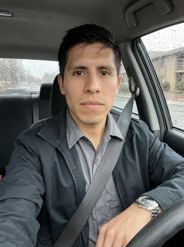 Benjamín, AI character: parked in his older compact car outside his apartment complex before work, rain dots on the windshield and…