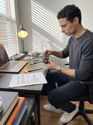 Matheus, AI character: his apartment desk covered with calipers, a laptop, job notes, and a small carburetor part from the Civic