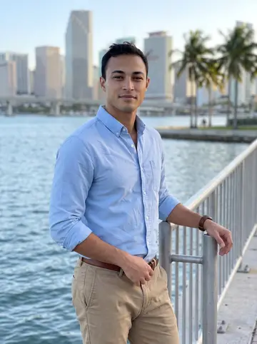 Armando, AI character: Biscayne Bay waterfront at dusk, leaning lightly on the metal railing with downtown Miami soft in the distance
