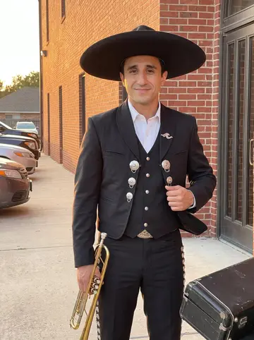 Ulises, AI character: outside a Houston wedding hall after a Saturday mariachi set, warm brick wall and parked cars behind him