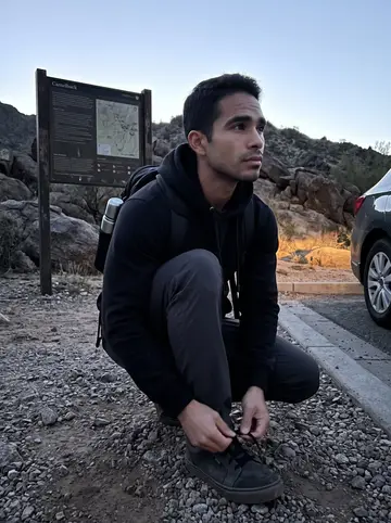 Alejandro, AI character: Camelback Mountain trailhead in Phoenix before sunrise, pale desert rocks and a parked car behind him