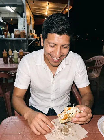 Ismael, AI character: a crowded Chula Vista taco stand at night, plastic tables and salsa bottles behind him