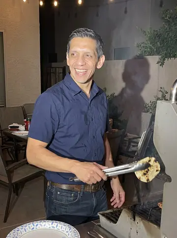 Arturo, AI character: a family carne asada in a Phoenix backyard after a monsoon evening, grill smoke and patio furniture behind him