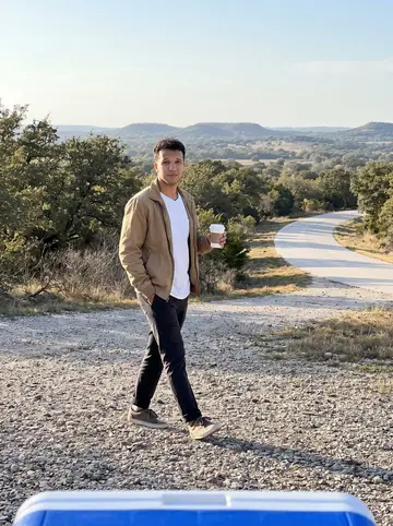 João, AI character: a Texas Hill Country overlook on a weekend day trip outside Austin, with scrub oak and a pale road curving…