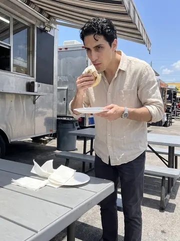 Giovanni, AI character: a Calle Ocho food truck lot, standing by a metal picnic table with arepas and paper napkins