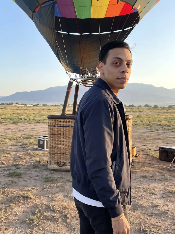 Thales, AI character: the west-side Albuquerque balloon launch field at dawn, wicker basket and a half-inflated balloon behind…