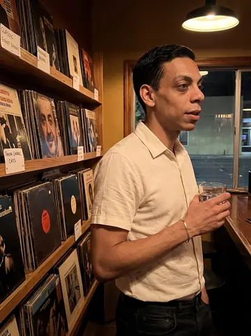 Thales, AI character: a small Albuquerque bar during a bossa nova vinyl night, standing near a wall shelf of records and dim…