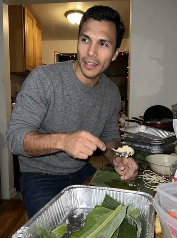 Félix, AI character: his mother's Bronx apartment kitchen during a December pasteles assembly line, aluminum trays and banana…
