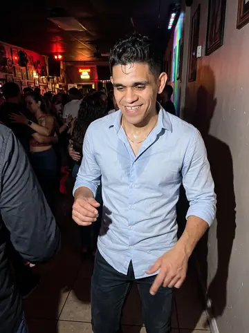 Yadiel, AI character: small salsa club in Hialeah after work, crowded dance floor with colored wall lights behind him