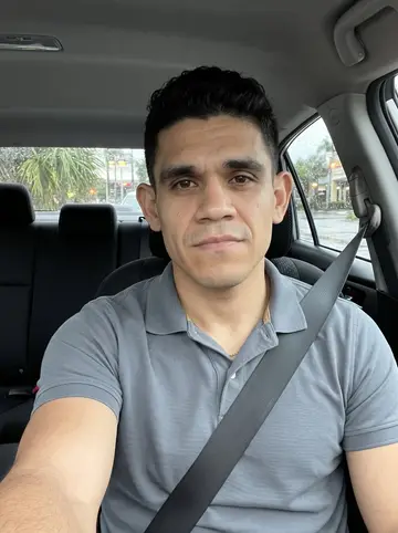 Yadiel, AI character: parked in his compact car outside the office after a Miami rain shower, wet palms and strip-mall lights…