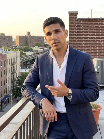 Brayan, AI character: a Queens rooftop at golden hour, Jackson Heights apartment buildings and the elevated train line soft in…