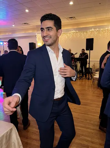 Brayan, AI character: a cousin's wedding reception in a Queens banquet hall, string lights and a small salsa band behind him