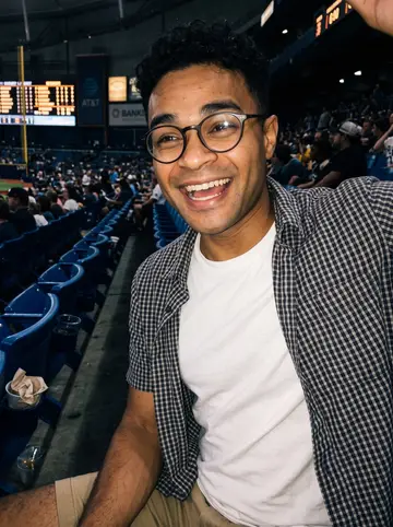 Raydel, AI character: cheap seats at a Tampa Bay Rays game, plastic cupholders and blue stadium seats around him