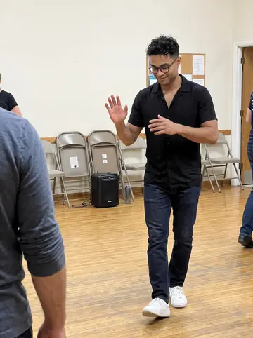 Raydel, AI character: a community salsa class in a church hall, scuffed wood floor and folding chairs along the wall
