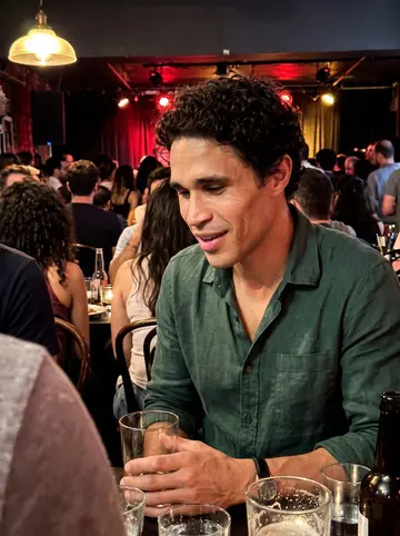 Vinícius, AI character: crowded Lower East Side Brazilian music night, small stage lights behind him and people packed close…