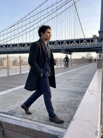 Vinícius, AI character: Brooklyn Bridge walkway on a clear cold morning before work, the phone propped low on a bench rail