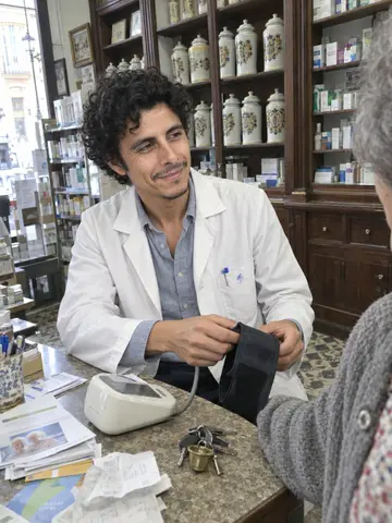 Javier, AI character: inside his small old corner pharmacy in Cádiz, wooden shelves with a row of antique ceramic pharmacy jars,…