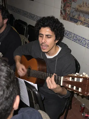 Javier, AI character: a cramped carnival peña rehearsal room in Cádiz, tiled walls, folding chairs, old chirigota posters, other…
