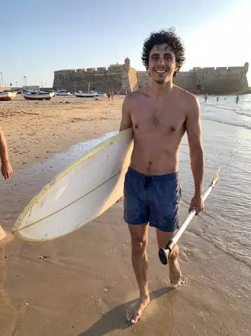 Javier, AI character: La Caleta beach in Cádiz at sunset, wet sand, the old stone castle and fishing boats behind him, a few…