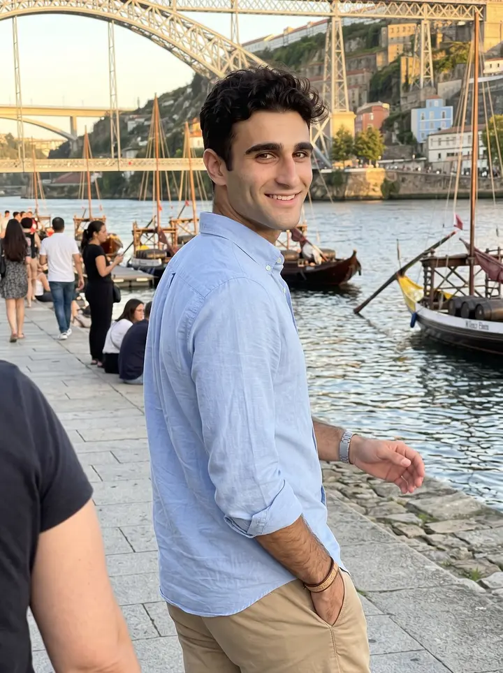 Vasco, AI character: the Ribeira waterfront in Porto near the Douro, rabelo boats behind him and the Dom Luís bridge catching…