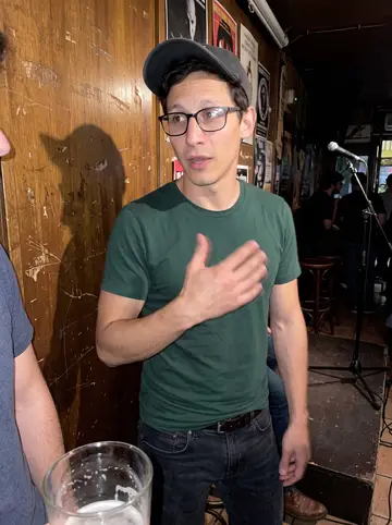 Pelayo, AI character: a small open mic bar in Malasaña after his set, standing beside a narrow stage and a scratched wooden wall