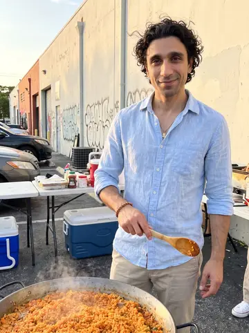 Denis, AI character: a Little Havana parking lot on a Sunday evening, beside a wide paella pan set over an open gas burner,…