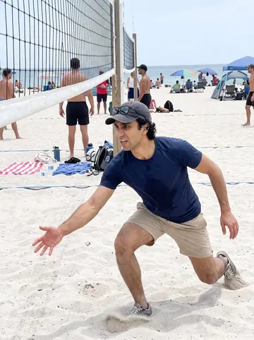 Denis, AI character: a public beach volleyball court on Miami Beach, sand bright under midday sun, friends and a net behind him