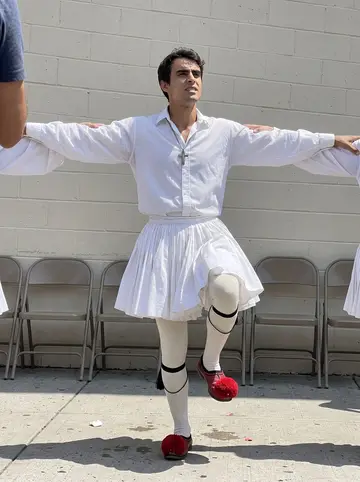 Napoleon, AI character: Astoria Greek street festival on a bright summer Saturday, painted church hall wall and folding chairs in…