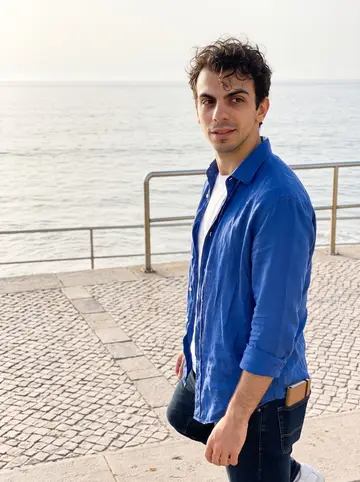 Vítor, AI character: Carcavelos boardwalk just after an early Atlantic swim, low railings and pale water behind him