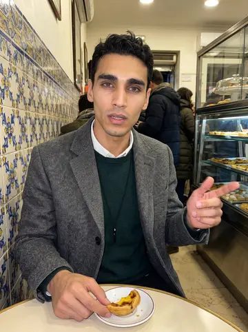 Telmo, AI character: inside a crowded old Lisbon pastry shop at night, tiled walls and glass counter behind him
