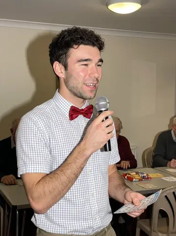 Darnell, AI character: the care home common room on Sunday bingo night, folding tables, prize biscuits and paper cards behind him