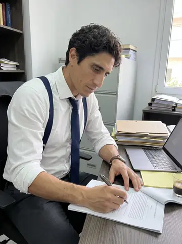 Theofilos, AI character: his commercial litigation office in Athens, narrow desk stacked with case folders, laptop, legal pads, and…