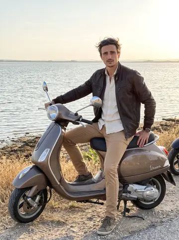 Federico, AI character: a roadside pullout near Albufera lake at sunset, his old Vespa-style motorbike parked beside dry summer grass